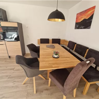 A modern dining area with a large wooden table and comfortable chairs. In the background, an elegant kitchen and a piece of artwork on the wall can be seen.