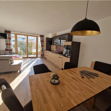 A modern living room with an open kitchen. A large wooden dining table and a cozy seating area by the window are clearly visible.