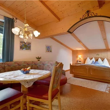 A cozy interior with a dining table and a bedroom in the background. The wooden beam ceiling and the large window front create a friendly atmosphere.