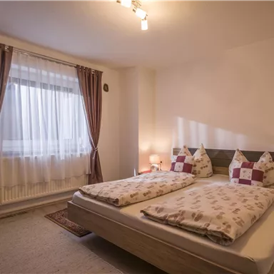 A cozy bedroom with two beds, light curtains, and a window. There is soft lighting and decorative pillows.