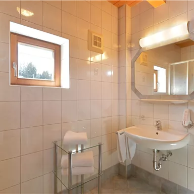 Ein modernes Badezimmer mit hellen Fliesen und einem Waschbecken. Es gibt ein Fenster, das Tageslicht hereinlässt, sowie Handtücher auf einem Glastisch.