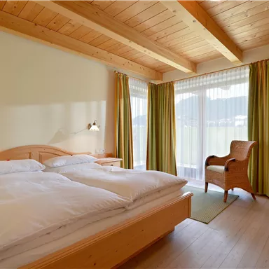 Ein helles Schlafzimmer mit einem Doppelbett und Holzdecke. Der Raum hat große Fenster mit Vorhängen und einen bequemen Sessel.