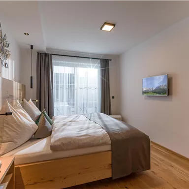 Ein modernes Schlafzimmer mit einem großen Bett und Holzmöbeln. Vor dem Fenster gibt es Vorhänge und einen Fernseher an der Wand.