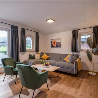 Ein modernes Wohnzimmer mit einem grauen Sofa und grünen Sesseln. Große Fenster bieten einen Blick auf die Natur.