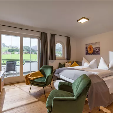 Ein helles Zimmer mit einem großen Bett, zwei gemütlichen Sesseln und einem Balkon, der einen schönen Ausblick auf die Landschaft bietet. Die Einrichtung ist modern und einladend.