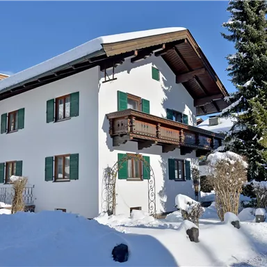 Ein hübsches Haus im Schnee mit grünen Fensterläden. Umgeben von schneebedeckten Bäumen und klarem Himmel.