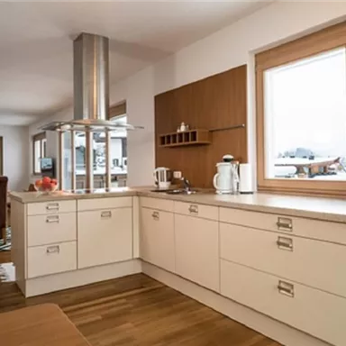 Eine moderne, helle Küche mit Holzmöbeln und großen Fenstern. Im Hintergrund sieht man einen gemütlichen Wohnbereich mit einem Hinweisschrank.