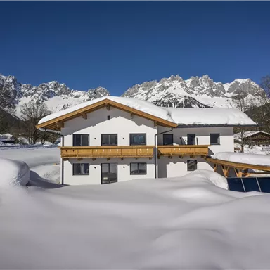 A modern house in a snowy environment with majestic mountains in the background. The sky is clear and blue.