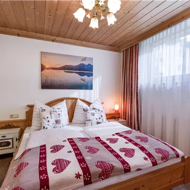 A cozy bedroom with a double bed and a wooden ceiling. The room is bright with a window, curtains, and a beautiful wall mural.