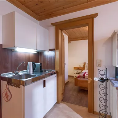 A small, bright kitchen with modern furniture and a wooden ceiling. The view leads to an adjacent room with a cozy bed.