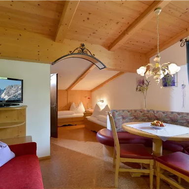 A cozy living room with wooden walls and a large window. A dining area with a table and chairs, as well as a television, are clearly visible.