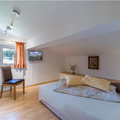 Ein gemütliches Zimmer mit einem niedrigen Dach und Holzfußboden. Es enthält ein Bett, einen Stuhl und einen Fernseher an der Wand.