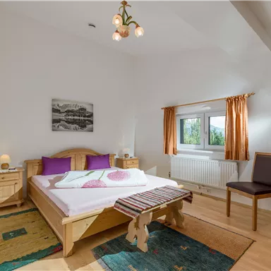 Ein gemütliches Schlafzimmer mit einem großen Bett und lila Kissen. Das Zimmer hat Holzfußboden, einen Stuhl und eine Fensteransicht.