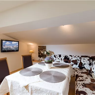 Ein gemütlicher Wohnraum mit einer beigen Couch im Kuhfell-Design und einem Essbereich. An der Wand hängt ein Fernseher, und ein Tisch ist mit einem weißen Tuch gedeckt.