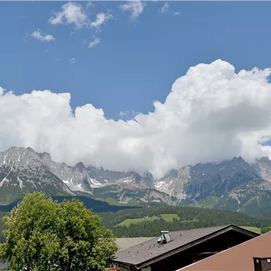 An impressive mountain landscape with large, wooded slopes and snow-capped peaks. The sky is blue, and white clouds are passing by.