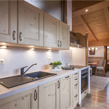 A modern kitchen with wooden cabinets and a bright countertop. In the background, a cozy living area is visible.