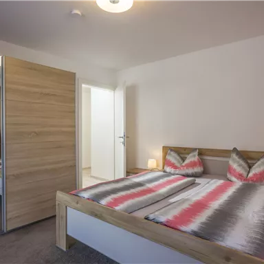 A cozy bedroom with a double bed and stylish pillows. There is a wooden wardrobe and plenty of natural light coming through the window.