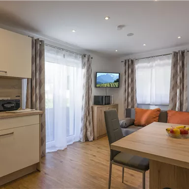 A bright and modern apartment with an open kitchen and a cozy living area. The space is furnished with a dining table, comfortable chairs, and a television.