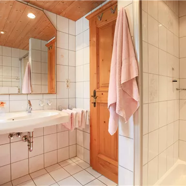 Ein helles Badezimmer mit gefliesten Wänden und einem großen Spiegel. Zu sehen sind ein Waschbecken, eine Dusche und ein Holzofen.