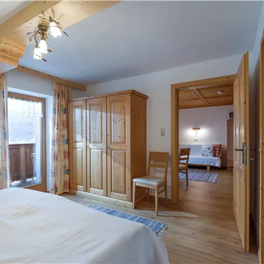 A cozy bedroom with a large bed and wooden details. A window with curtains and an open door lead to another room.