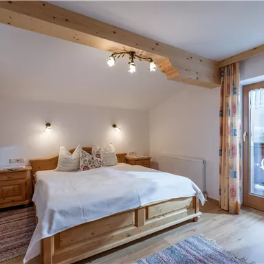 A cozy bedroom with a large bed and wooden furniture. The room has a balcony with a view and feels bright and inviting.