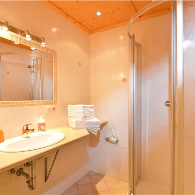 Ein modernes Badezimmer mit Dusche, Waschbecken und Spiegel. Weiße Handtücher sind ordentlich gestapelt auf einer Ablage.