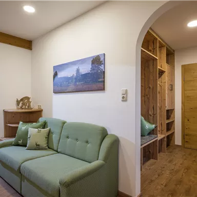 A cozy living space with a green sofa and warm light. A picture of a landscape hangs on the wall, and there is a wooden shelf on the right side.