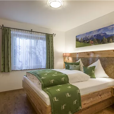 A cozy bedroom with a wooden bed and green bedcovers. The room has large windows and a beautiful wall decoration featuring a mountain landscape.