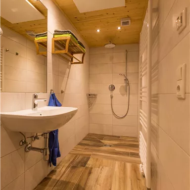 Ein modernes Badezimmer mit einer Dusche und einem Waschbecken. Der Boden ist aus Holzoptik, und es gibt Regale für Handtücher.