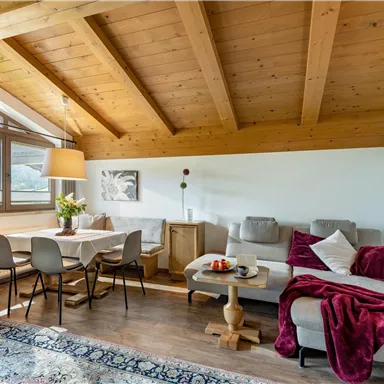 A cozy living room with wooden beams and large windows. The room is decorated with an elegant sofa, a table, and a warm blanket.