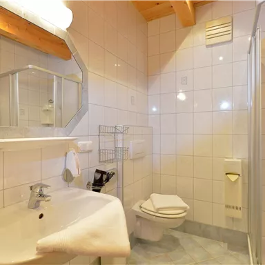 Ein modernes Badezimmer mit weißen Fliesen und Holzdetails. Es enthält eine Dusche, ein Waschbecken und eine Toilette.