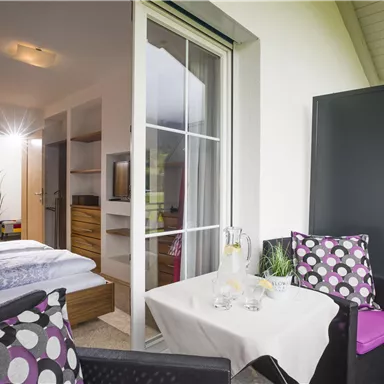 Ein gemütlicher Balkon mit einem kleinen Tisch und zwei Stühlen. Im Hintergrund sieht man ein helles Zimmer mit einem modernen Einrichtungsstil.