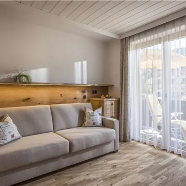 A modern living room with a beige sofa and a wooden table. Large windows with curtains offer a view of a balcony.