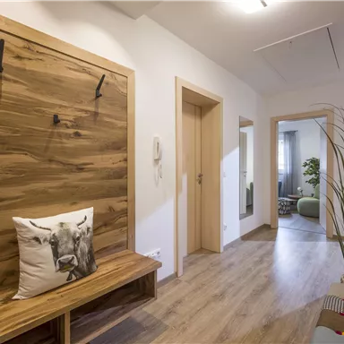 Ein heller Flur mit Holzmöbeln und einer gemütlichen Bank. An der Wand hängen einige Haken, und es gibt einen kleinen Pflanzenakzent.