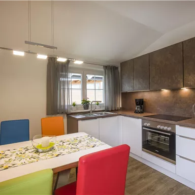 Eine moderne Küche mit hellen Schränken und einem kleinen Esstisch. Bunte Stühle und Fenster sorgen für ein einladendes Ambiente.