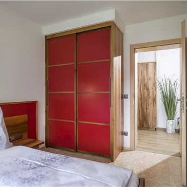 Ein gemütliches Schlafzimmer mit einem schlichten Bett und einem roten Kleiderschrank. Im Hintergrund ist ein heller Eingangsbereich mit einer Pflanze sichtbar.
