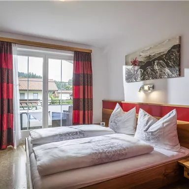 Ein gemütliches Schlafzimmer mit einem Doppelbett und modernen Möbeln. Große Fenster mit roten Vorhängen bieten einen Blick nach draußen.