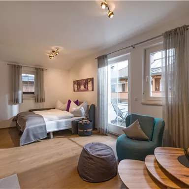 Ein gemütliches Zimmer mit einem Doppelbett und Platz zum Sitzen. Große Fenster sorgen für viel Tageslicht und eine angenehme Atmosphäre.