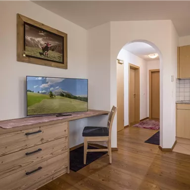 Ein modernes Zimmer mit Holzfußboden und einer Kommode. Im Raum befindet sich ein Fernseher, ein Tisch mit Stuhl und eine kleine Küche.