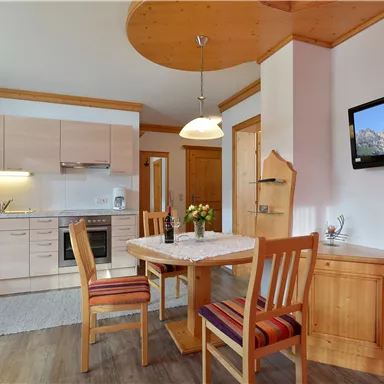 A modern kitchen with wooden details and a dining table. A television is mounted on the wall, and a vase of flowers sits on the table.