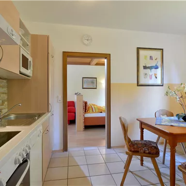 Eine helle Küche mit Holzschränken und einem Tisch. Im Hintergrund ist ein Wohnzimmer mit einem roten Sofa zu sehen.