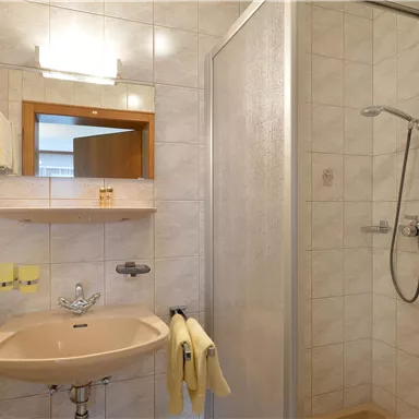 A bright bathroom with a sink, a mirror, and a shower. The walls are covered with light tiles.