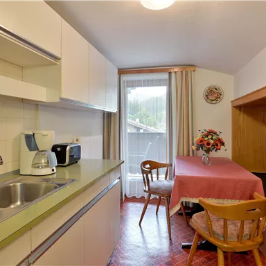 A bright kitchen with modern furniture, a dining area, and a window that lets in daylight. On the table are flowers and fresh fruit.