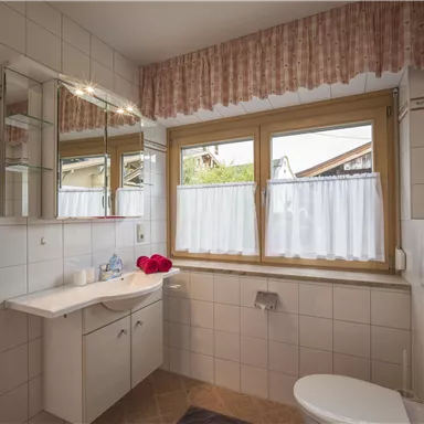 Ein helles Badezimmer mit weißen Fliesen und einem großen Fenster. Rote Handtücher hängen an der Wand, und ein Waschbecken mit Spiegel ist gut sichtbar.