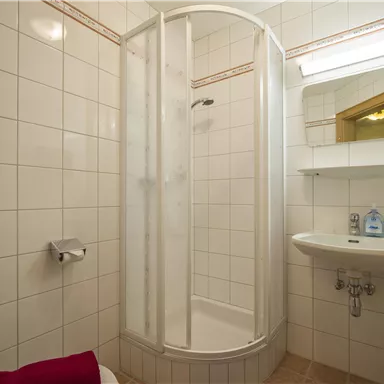 Ein kleines Badezimmer mit einer Duschkabine, einem Waschbecken und weißen Fliesen. Rote Handtücher sind als Akzent hinzugefügt.