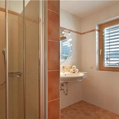 A modern bathroom with a shower and a sink. It features large windows with wooden blinds and a cozy atmosphere.