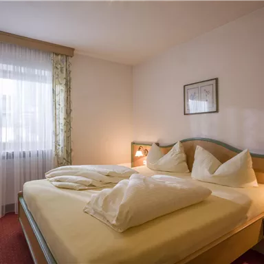 Ein gemütliches Schlafzimmer mit einem großen Bett und zwei Kissen. Die Fenster sind mit Vorhängen geschmückt und bieten Tageslicht.