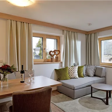 Ein gemütlicher Wohnraum mit einem grauen Sofa und einem Holztisch. Große Fenster lassen viel Tageslicht herein und schaffen eine einladende Atmosphäre.