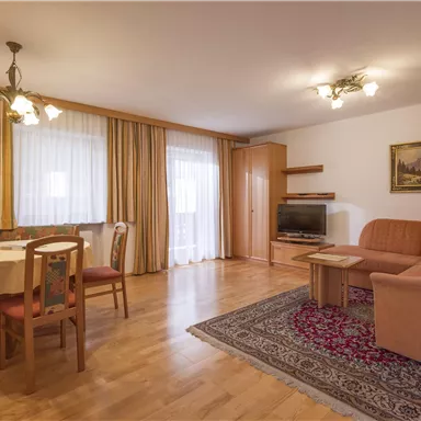 Ein gemütliches Wohnzimmer mit einem Tisch, Stühlen und einer Couch. Große Fenster lassen viel Tageslicht herein und der Boden ist aus Holz.