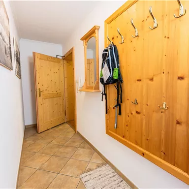 Ein heller Flur mit einem Holzkleiderhaken und einem Rucksack. Die Wände sind in neutralen Farben gehalten, und eine Haustür führt nach draußen.